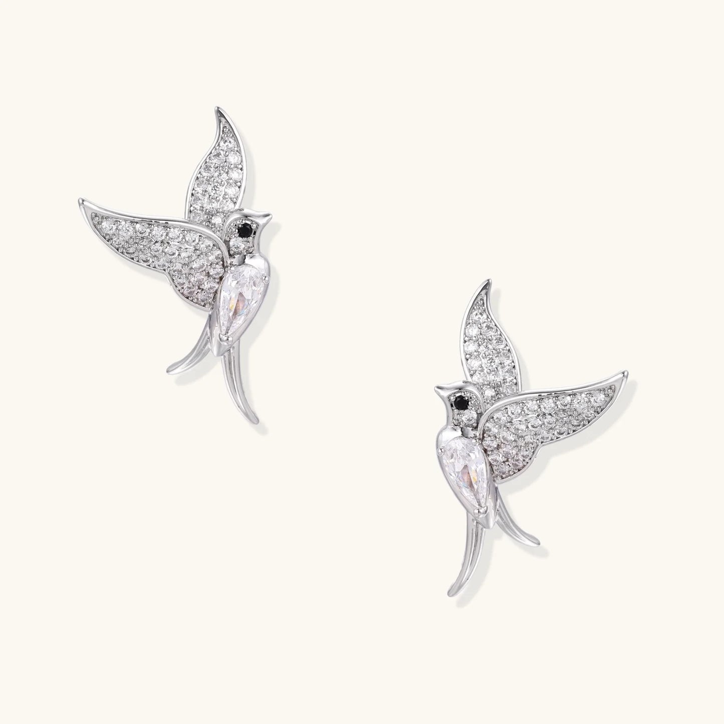 Pair of white gold-plated hummingbird stud earrings with pavé zirconia stones and teardrop crystal bodies on cream background