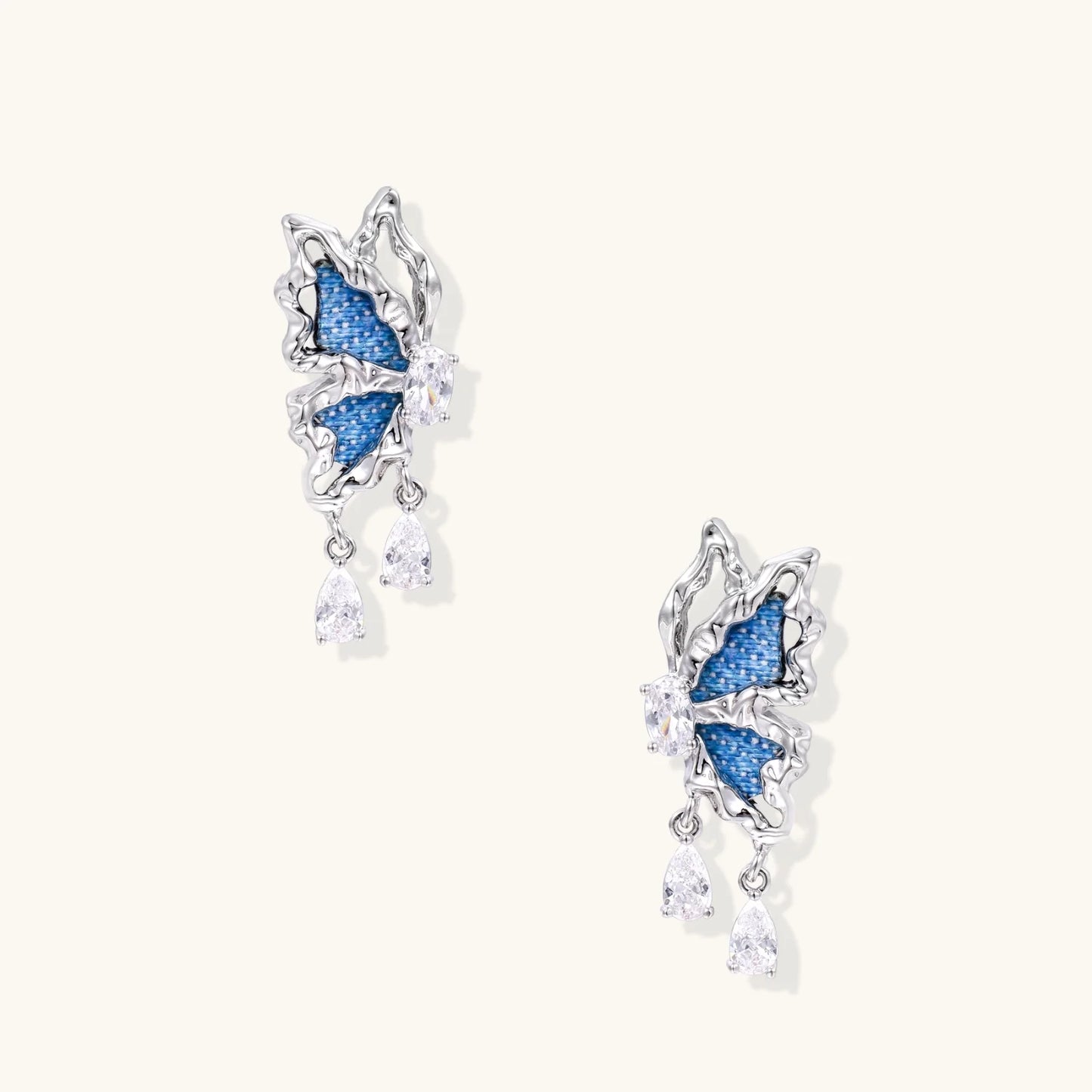 Silver butterfly earrings with blue denim fabric wings and dangling teardrop crystals on cream background