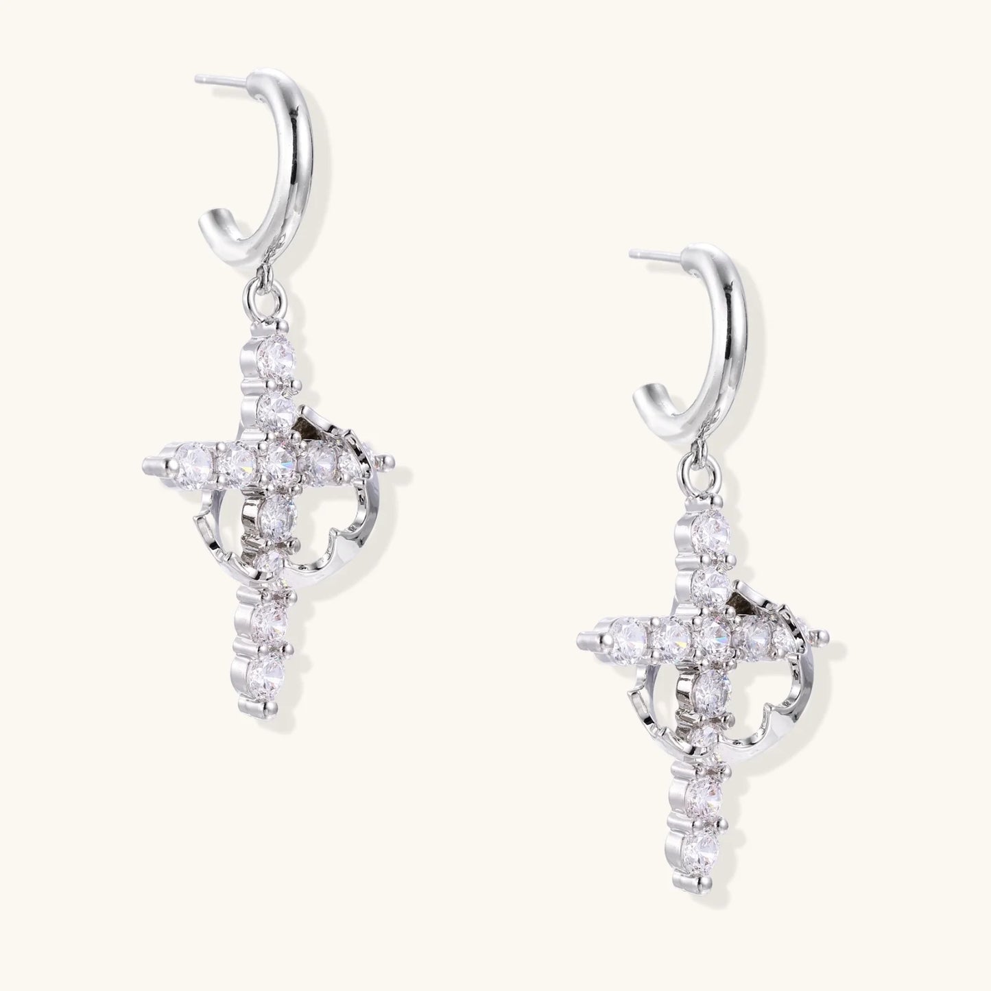 White gold-plated hoop earrings with sparkling zirconia cross charms dangling against cream background
