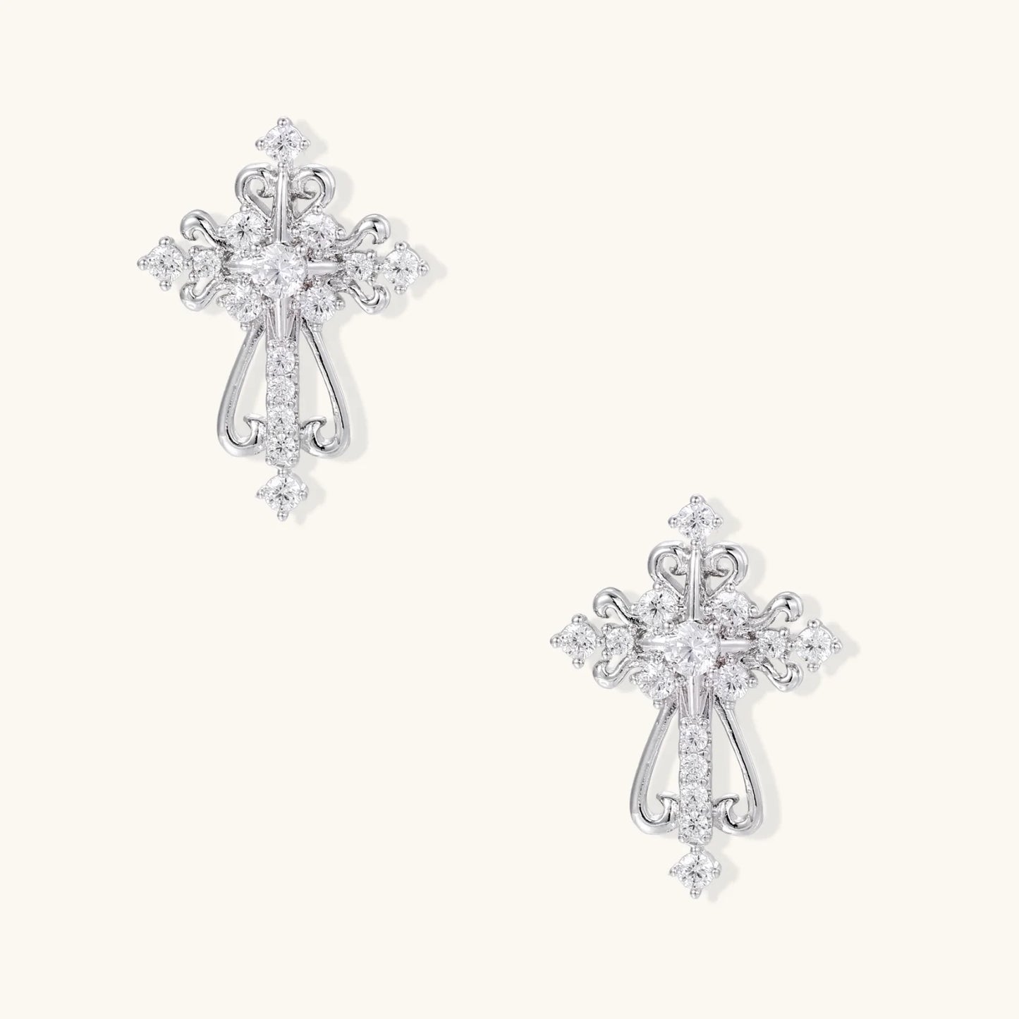 White gold-plated baroque cross studs with zirconia stones and ornate scrollwork on cream background.