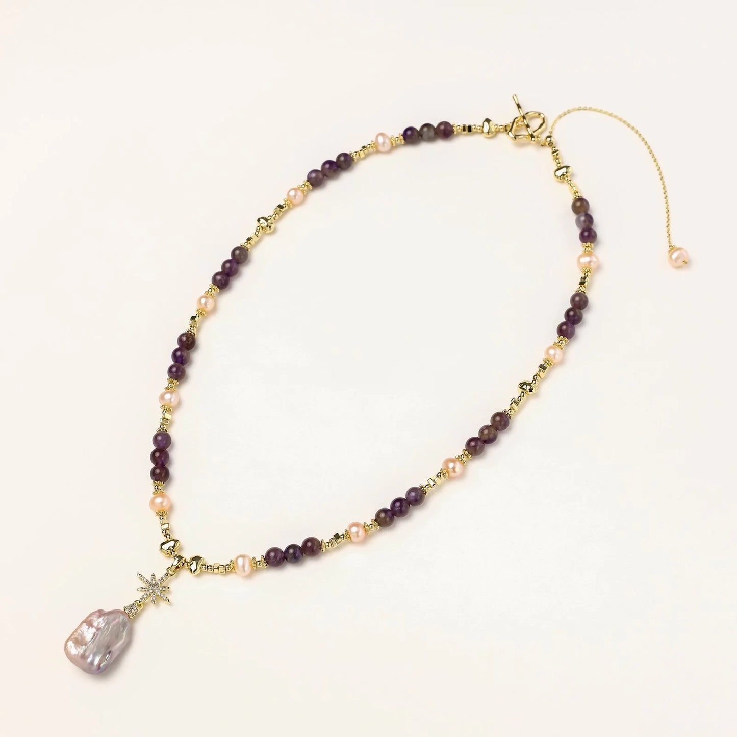 Gold necklace with iridescent baroque pearl pendant, purple amethyst beads, and crystal star charm on cream background