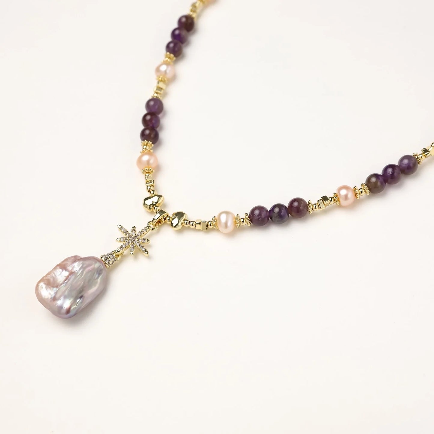 Gold necklace with iridescent baroque pearl pendant, purple amethyst beads, and crystal star charm on cream background