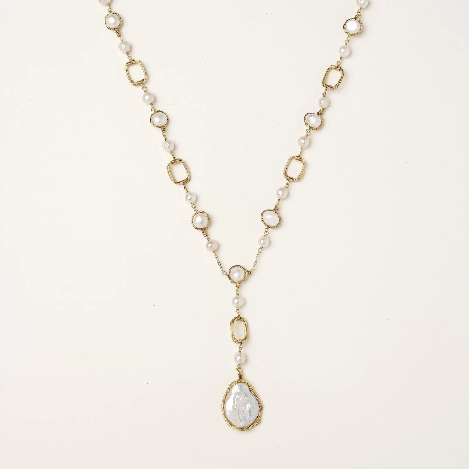 Y-shaped gold necklace with alternating geometric links, freshwater pearls, and baroque pearl pendant on cream background