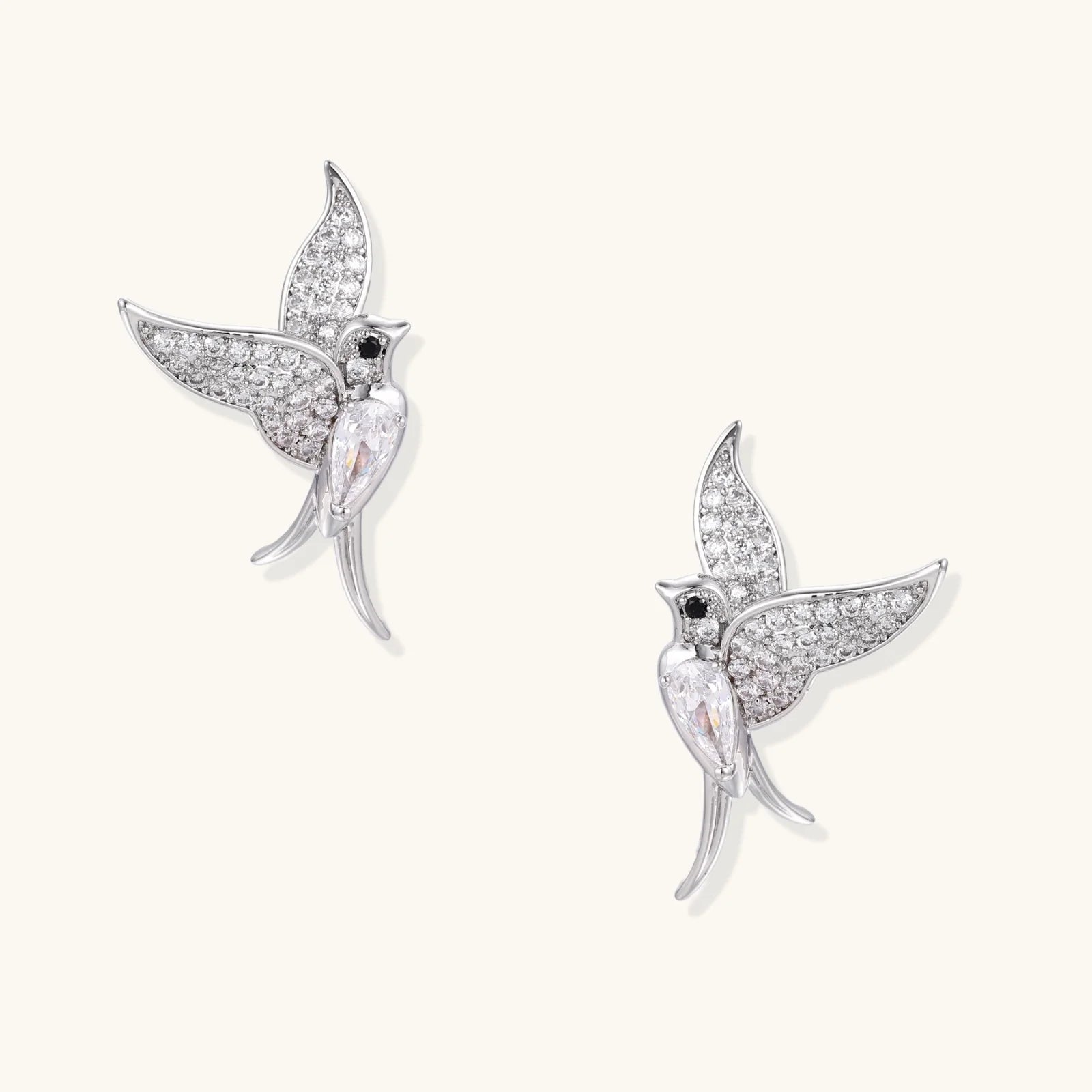 Pair of white gold-plated hummingbird stud earrings with pavé zirconia stones and teardrop crystal bodies on cream background
