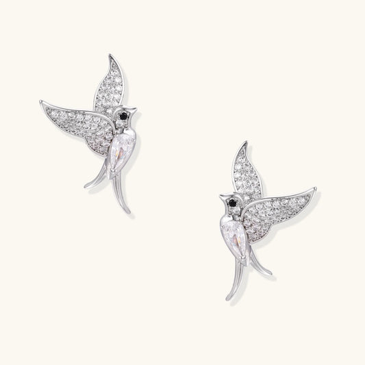 Pair of white gold-plated hummingbird stud earrings with pavé zirconia stones and teardrop crystal bodies on cream background