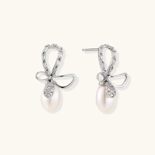 Silver bow-shaped stud earrings with baguette crystals and dangling white freshwater pearls on cream background