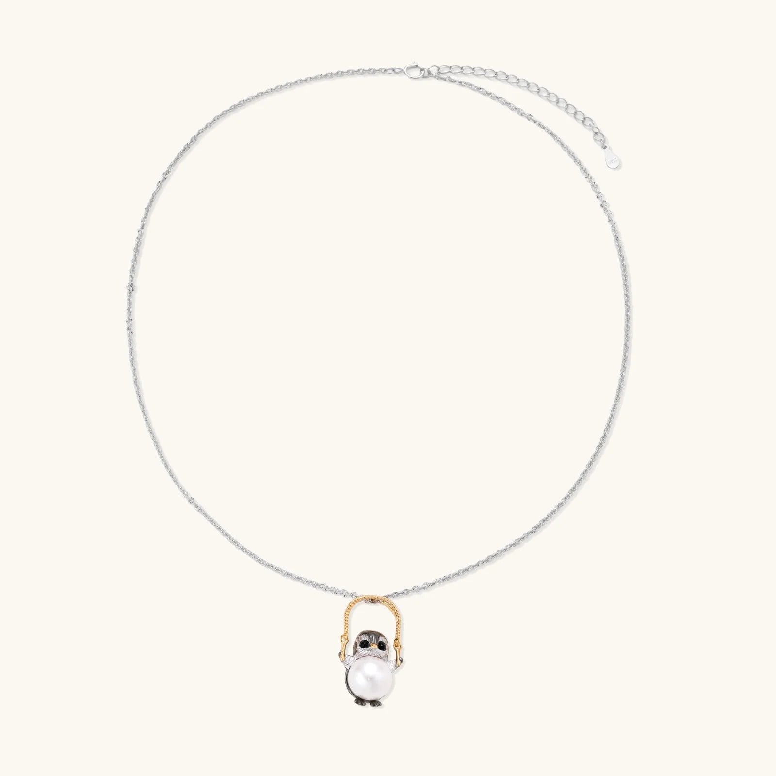 Whimsical owl pendant holding a white pearl on a gold swing, rhodium-plated silver chain on cream background.