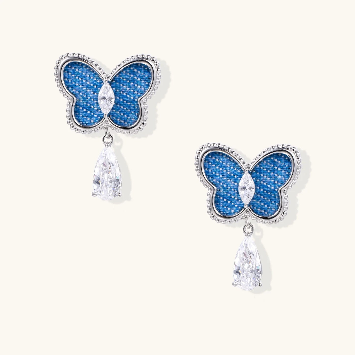Blue denim butterfly earrings with silver frame and teardrop crystal dangles on cream background