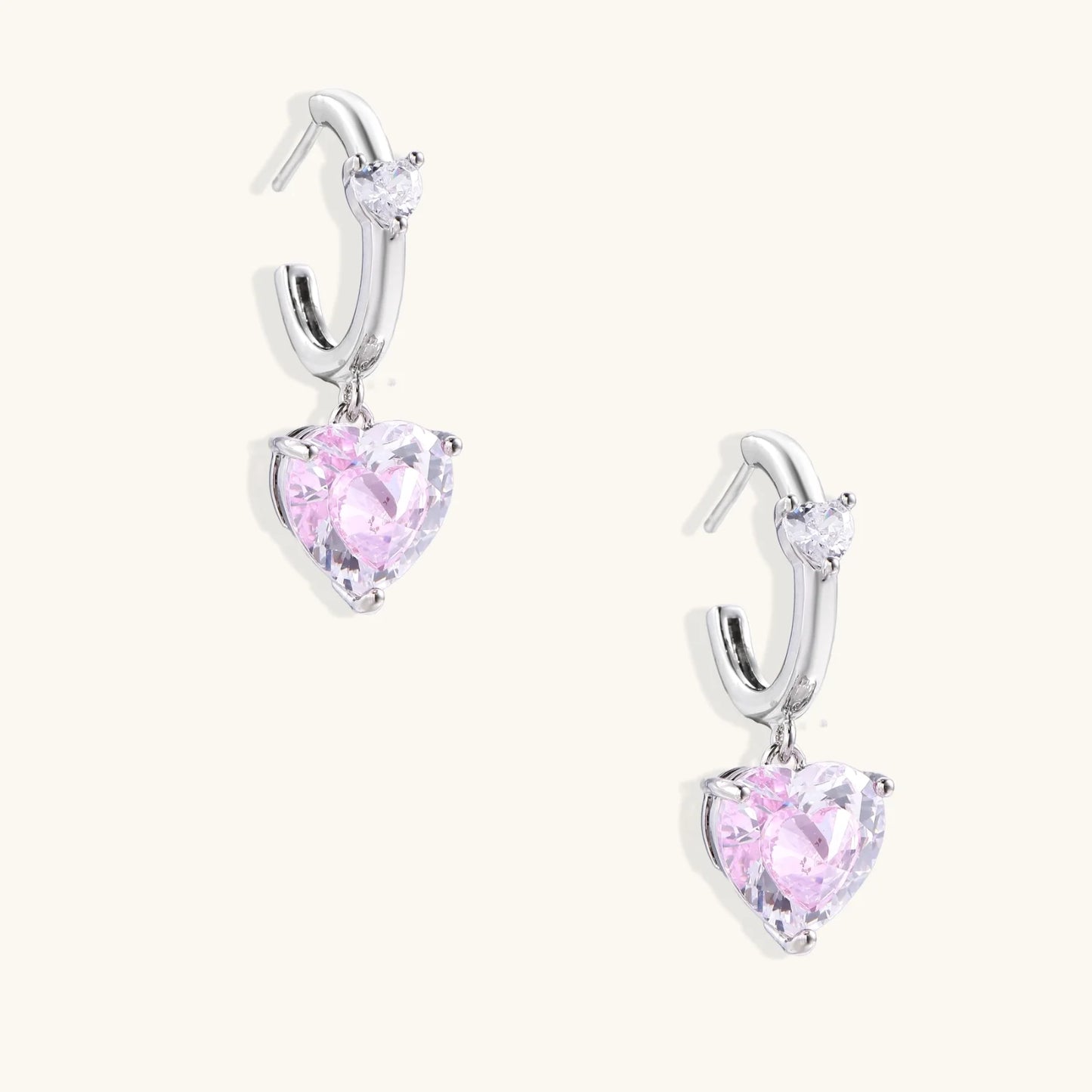 Silver huggie hoops with pink heart-cut zirconia pendants on white background, eco-friendly white gold plated brass