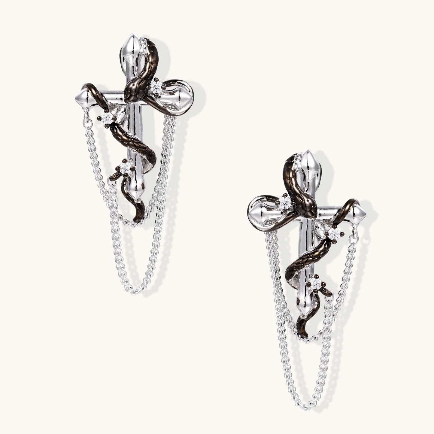 Silver dangle earrings with sculptural serpent coiled around cross shape and delicate chain accents on cream background