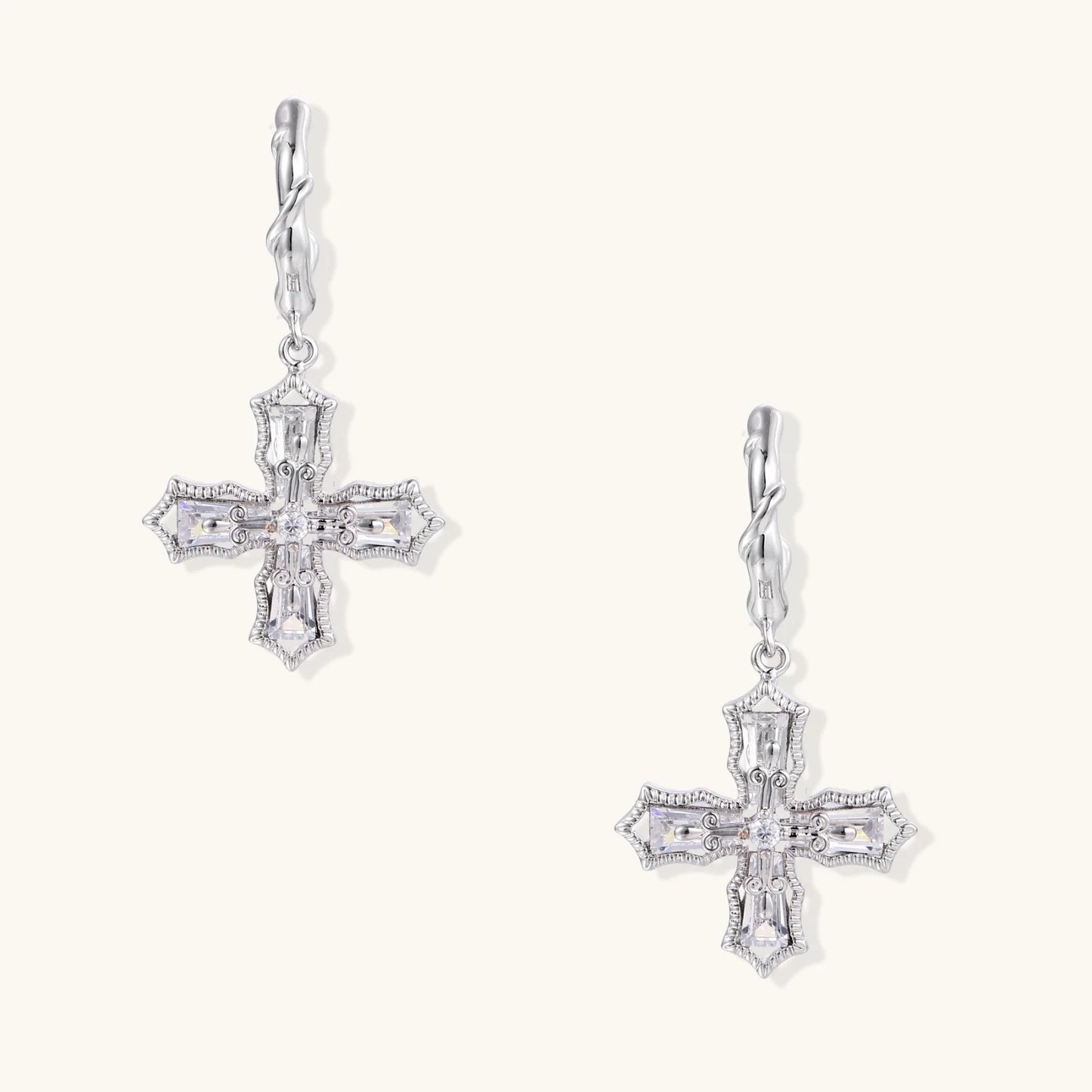 Ornate white gold-plated cross earrings with baguette-cut zirconia stones and twisted leverback hooks on beige background