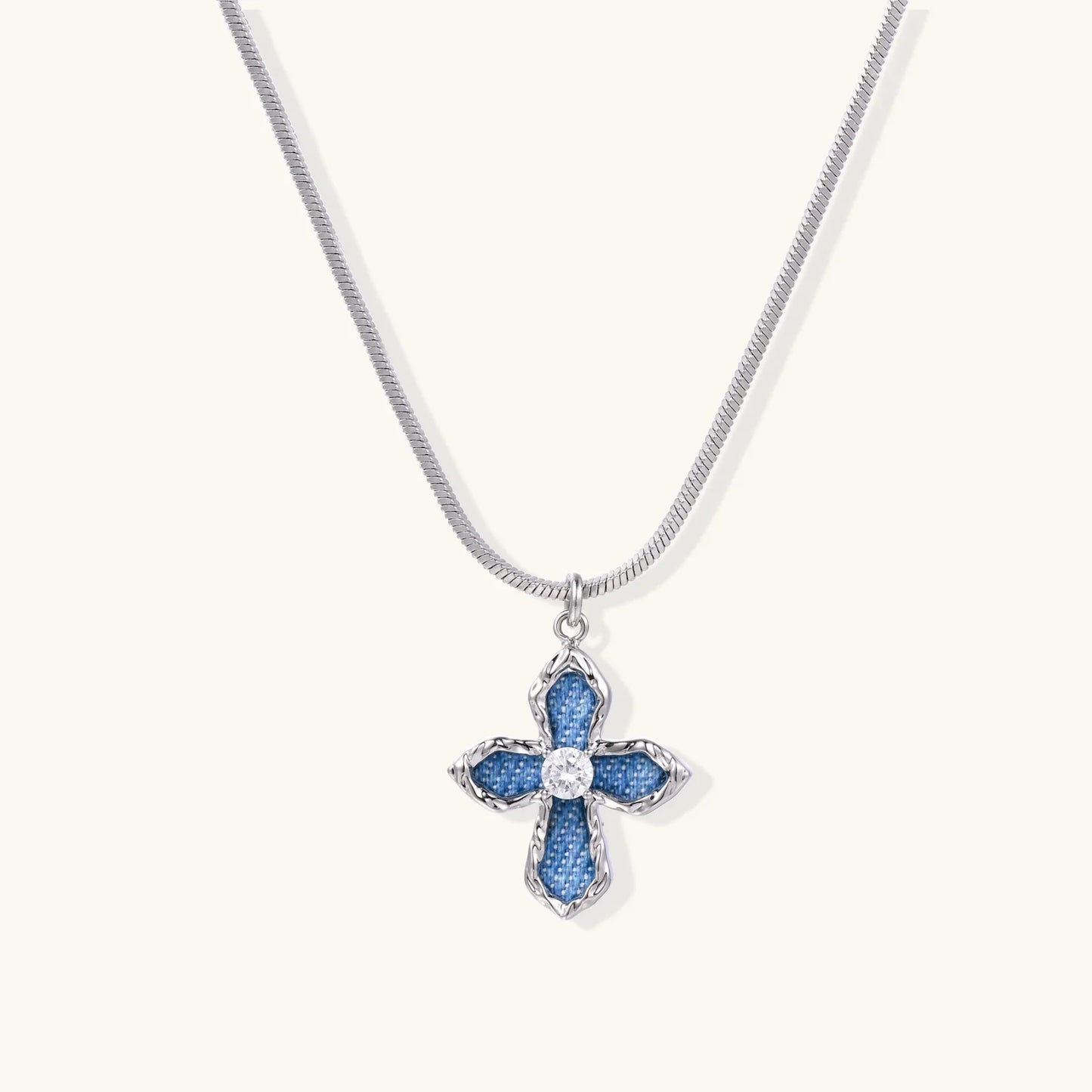 Blue denim-inlaid cross pendant with white gold frame and center zirconia on silver snake chain against cream background