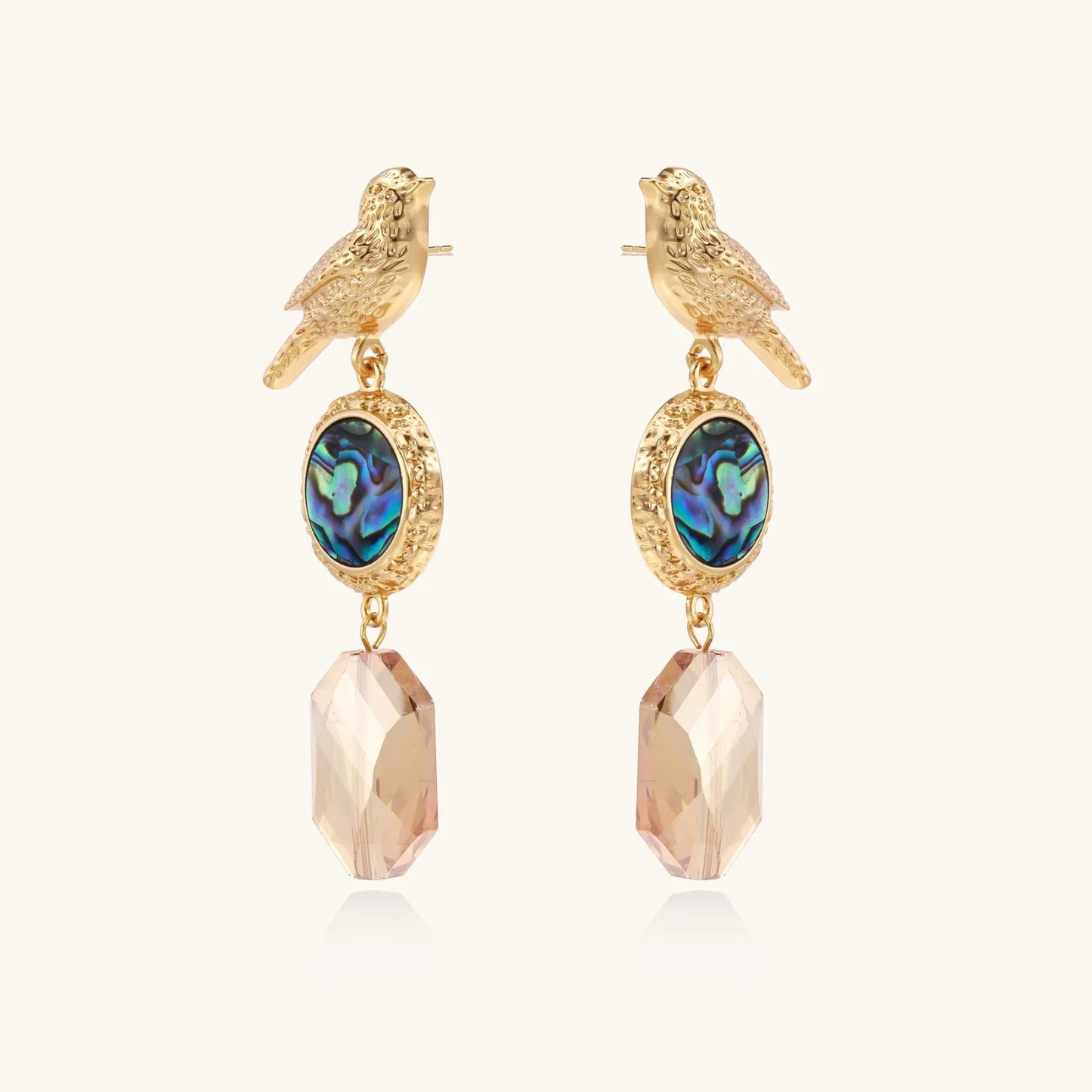 Gold-plated bird stud earrings with iridescent abalone shell medallions and champagne crystal drops on cream background