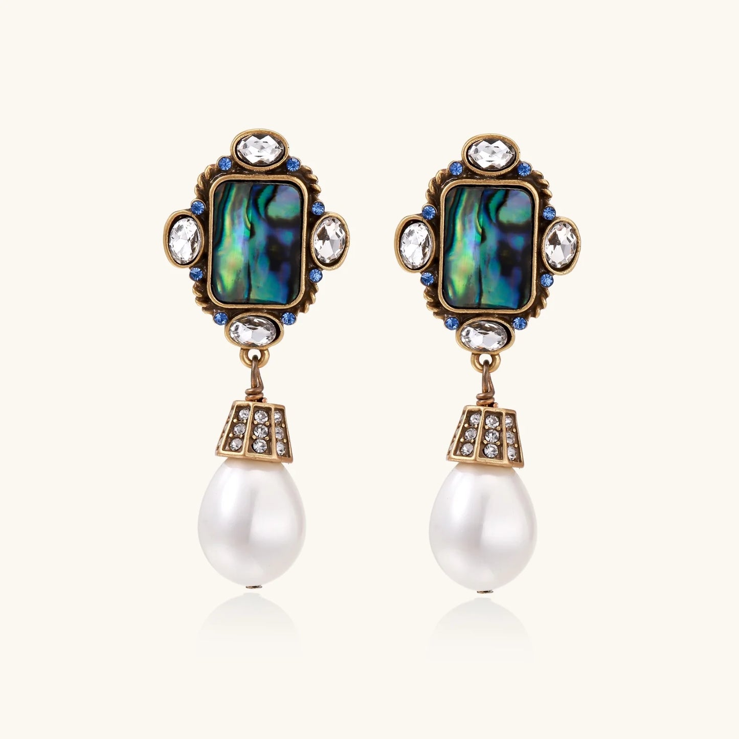 Ornate gold-plated earrings with iridescent abalone shell, crystals, blue stones, and white pearl drops on cream background.